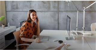 A young business professional seated at a desk in a modern office. She is smiling, looking at a computer monitor. She is wearing a striped blue and white button down shirt with a dark navy blazer. She is typing on a white computer keyboard with her right hand and holding a LAN line phone she is using with her left hand.