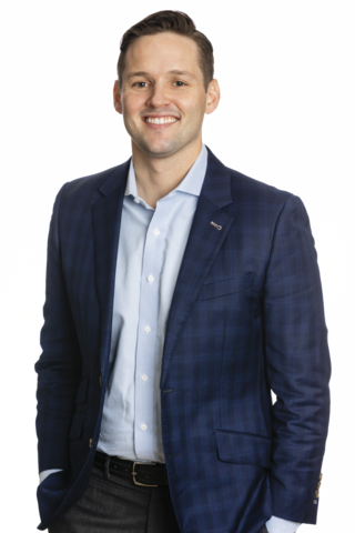 Michael is pictured smiling. He is wearing a dark blue sport coat, a light blue button down shirt, dark grey dress pants and a black leather belt. He is standing with his right hand tucked into his front pocket and his left arm resting at his side.