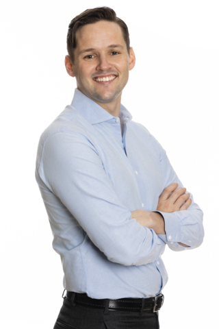 Michael is pictured smiling. He is wearing a light blue button down shirt, dark grey dress pants and a black leather belt. He is standing with his arms folded in front of him.