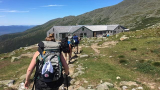 A group of women mountain climbing, carrying backpacks, walk in a single file line away from the camera, toward a grey structure at the summit of a mountain. The sun is shining, the grass on the mountain is green and it is summer. Beyond the structure, there are more mountains and the blue sky.