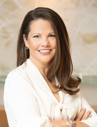 Amy is pictured smiling. She is wearing an ivory silk tie neck blouse. She is facing the camera while her body is angled slightly sideways and her arms are folded in front of her.