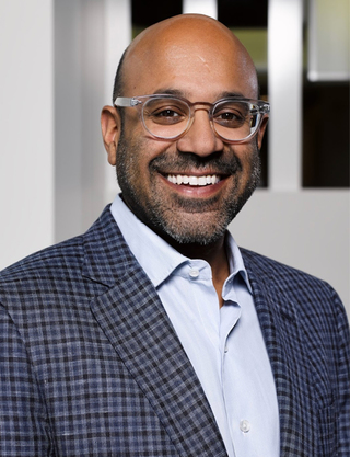 Niraj is pictured smiling and facing forward. He is wearing a dark teal sport coat with a plaid pattern, a light blue button down shirt and clear, acetate eyeglasses.