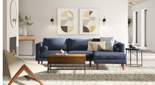 A modern living room is pictured, painted entirely in an ivory shade. In the center there is a mid-century modern sofa in slate blue cloth upholstery and two decorative pillows. Three tables surround the  sofa, one round iron side table to the left, one low wood and iron coffee table in the center and a wood and iron taller console table behind and to the right. Two pieces of wall art hang on the wall behind the sofa featuring round geometric shapes in neutral tones, with two black metal sconces on either side. To the right of the sofa is a a simple fireplace with only a round gold trimmed mirror hanging above.