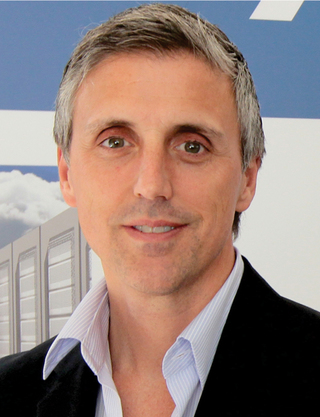 Chris is pictured smiling, facing forward. He is wearing a light blue button down shirt and a black sport coat.