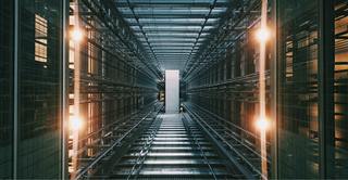 A long, dimly lit server room lined with grey-blue steel wracks. In the foreground, two vertically aligned lightbulbs give off a golden glow, on each side of the room.